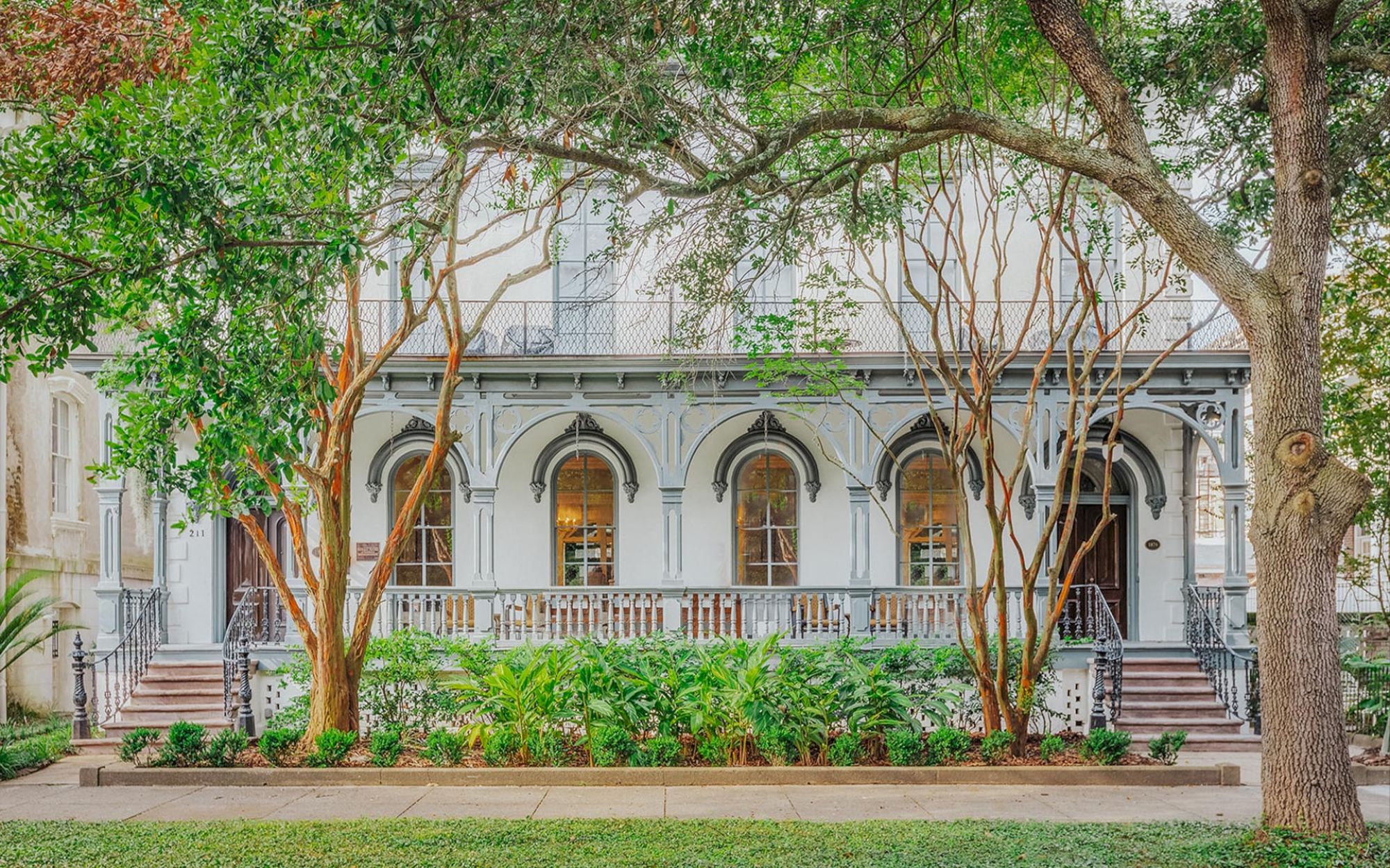 A classic, two-story building with arched windows and a porch is surrounded by lush greenery and trees on a sunny day, evoking a serene atmosphere.