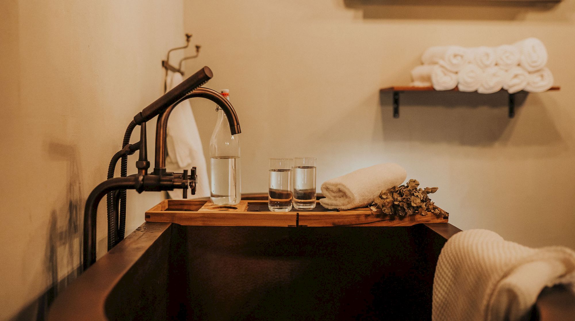 A cozy bathroom setup with a rustic faucet, wooden bathtub tray holding water glasses, folded towels, and a shelf with more towels.