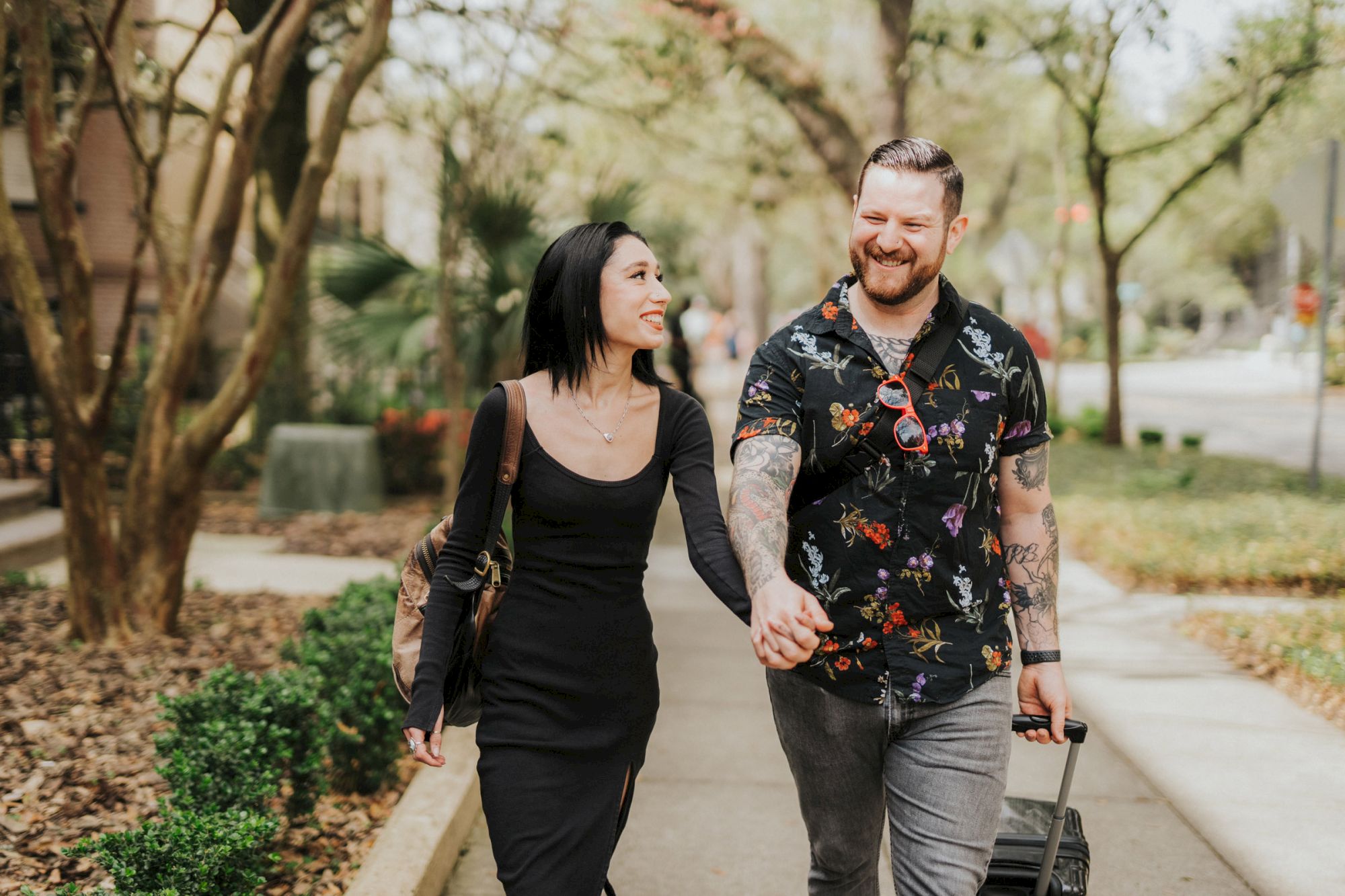 A couple is walking down a tree-lined sidewalk, holding hands and smiling, with the man pulling a suitcase and the woman carrying a backpack.