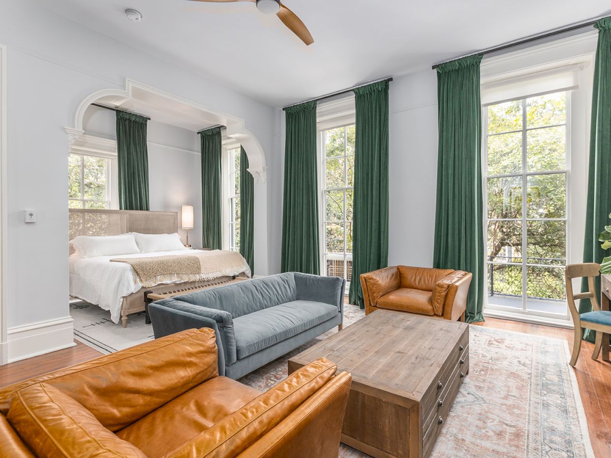 A spacious bedroom and sitting area features a bed, a gray sofa, brown leather chairs, green curtains, large windows, and a wooden coffee table.