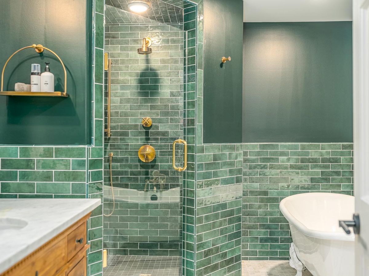 The image shows a bathroom with green tile walls, a glass shower enclosure with gold fixtures, a wooden vanity, and a white freestanding bathtub.