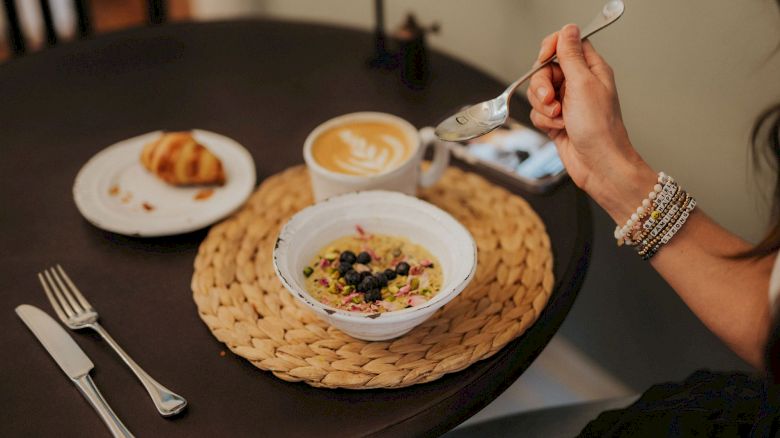 A person is having breakfast: oatmeal with berries, a croissant, and a latte on a table, with utensils on the side.