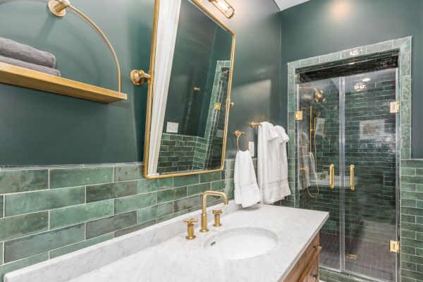 A modern bathroom with green tiles, a marble countertop, gold fixtures, a large mirror, and a walk-in glass shower with towels hanging.