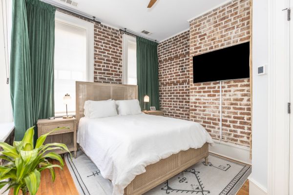 A cozy bedroom with a white bed, brick accent wall, TV, green curtains, bedside table, lamp, plant, and wooden floor, combining modern and rustic elements.