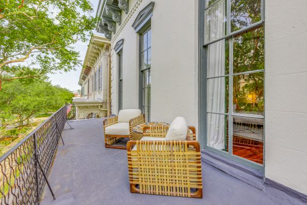 A balcony with two wicker chairs and cushions, iron railings, and a view of trees and neighboring buildings. The setting is serene and inviting.
