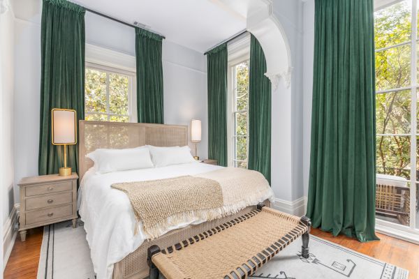 A cozy bedroom with green curtains, large windows, a double bed with neutral linens, bedside tables, and floor lamps on either side of the bed.