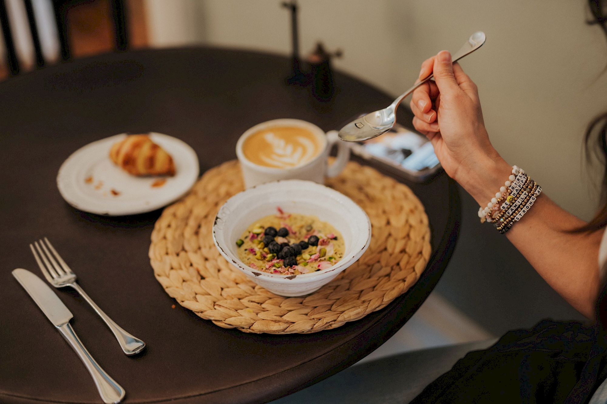 A person is having breakfast with a bowl of oatmeal topped with berries, a croissant, and a cup of coffee on a table with utensils.