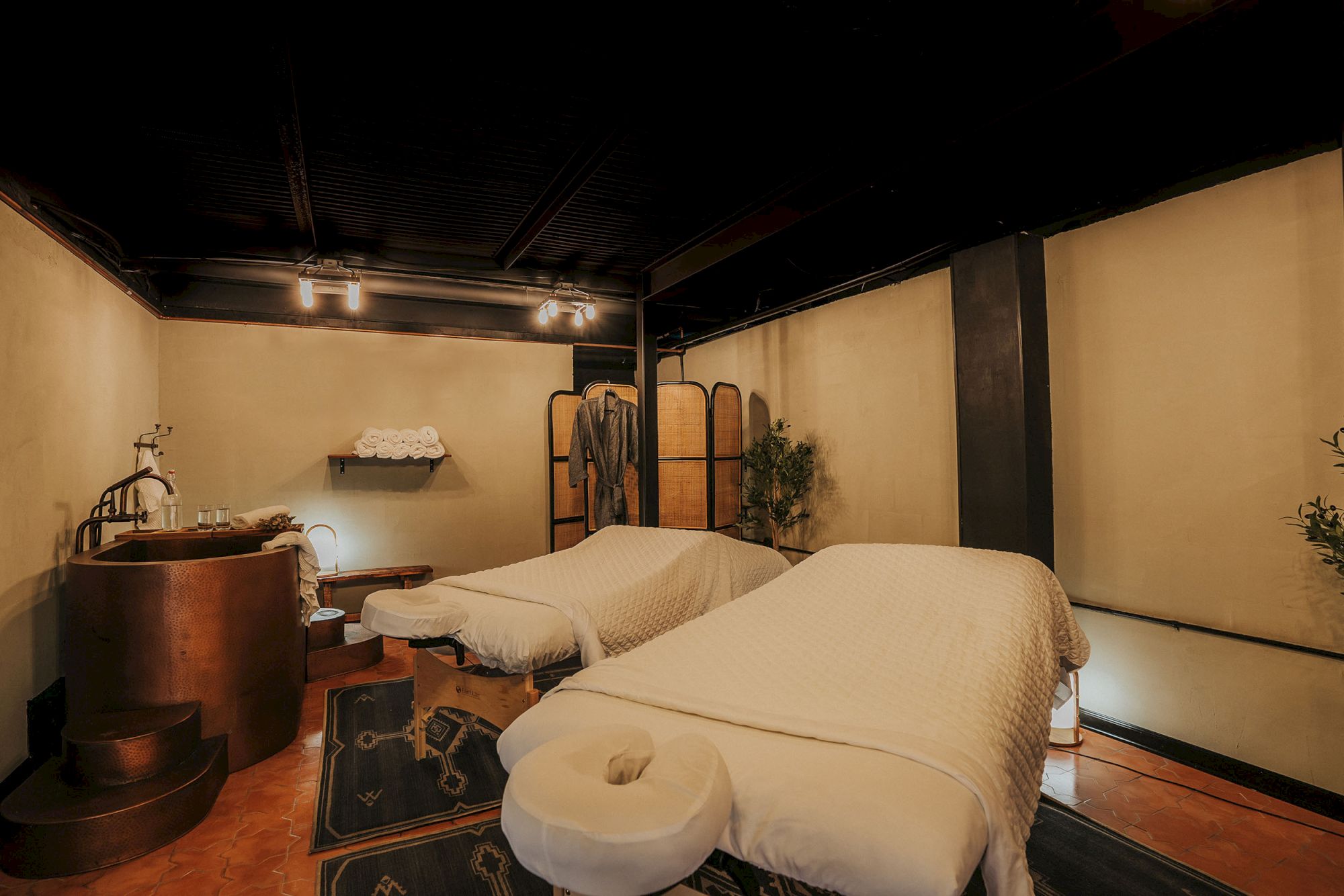 A serene spa room with two massage tables, towels, and a wooden tub nearby, creating a calming and inviting atmosphere for relaxation.