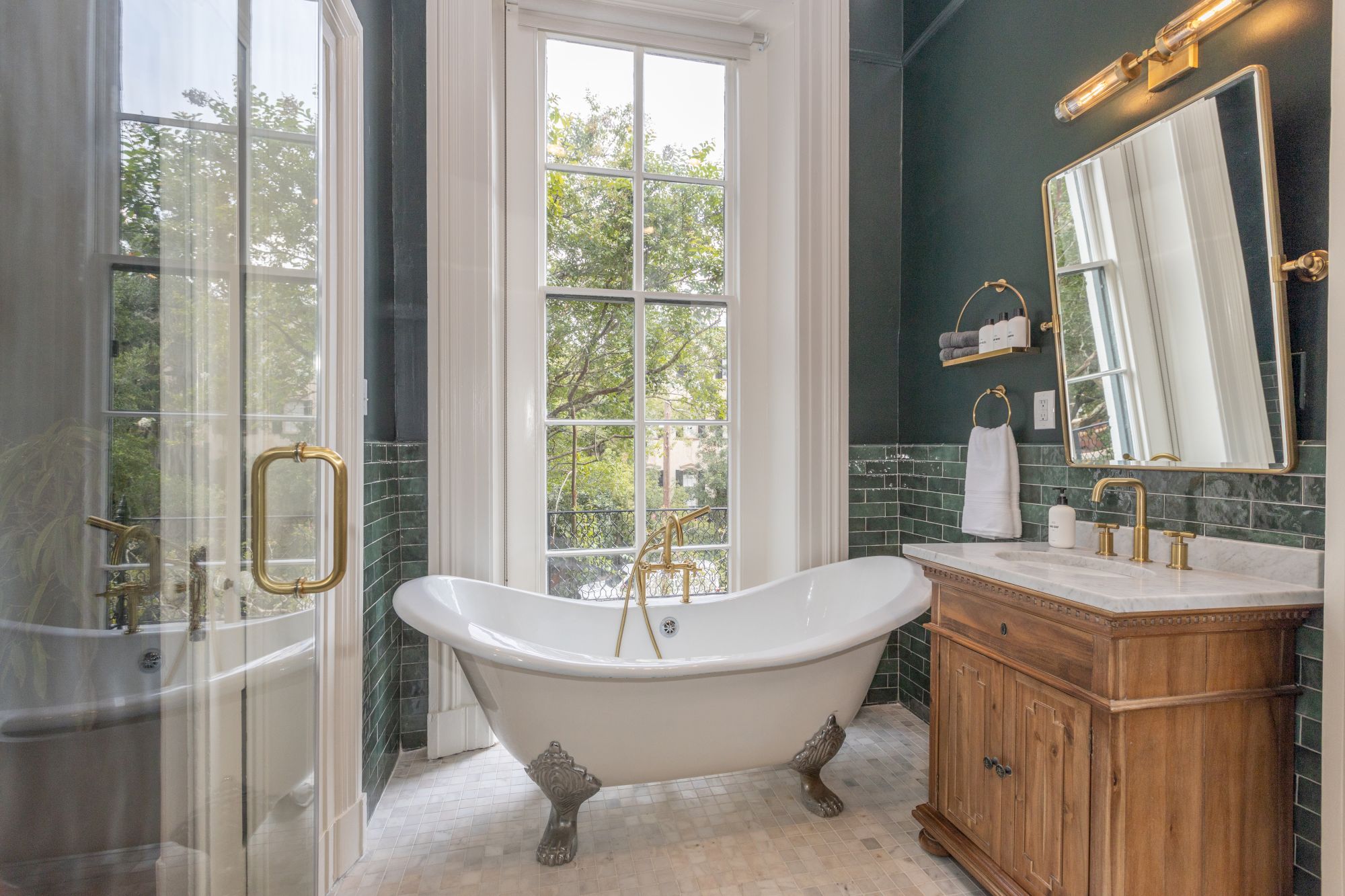 The image shows a bathroom with a central freestanding bathtub, a vanity with a sink and mirror, green tiled walls, and a large window providing natural light.