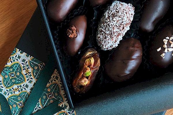 A box of assorted chocolate-covered dates with various toppings, placed on a decorative patterned cloth on a wooden surface.