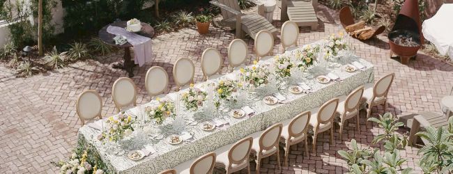 An outdoor dining area with a long table set for 12, decorated with floral arrangements and surrounded by chairs, in a garden setting.
