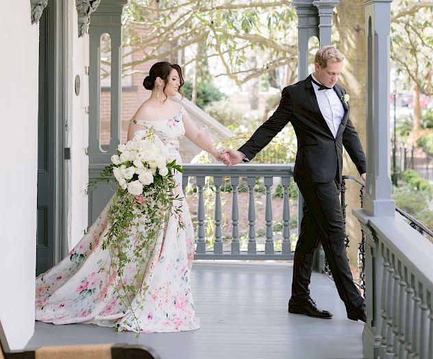 A couple dressed in formal attire, possibly a bride and groom, walk on a beautifully decorated porch with greenery in the background, holding hands.