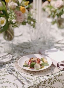 A beautifully set table with floral arrangements, forks/knives, and a plate of gourmet food on a patterned tablecloth.