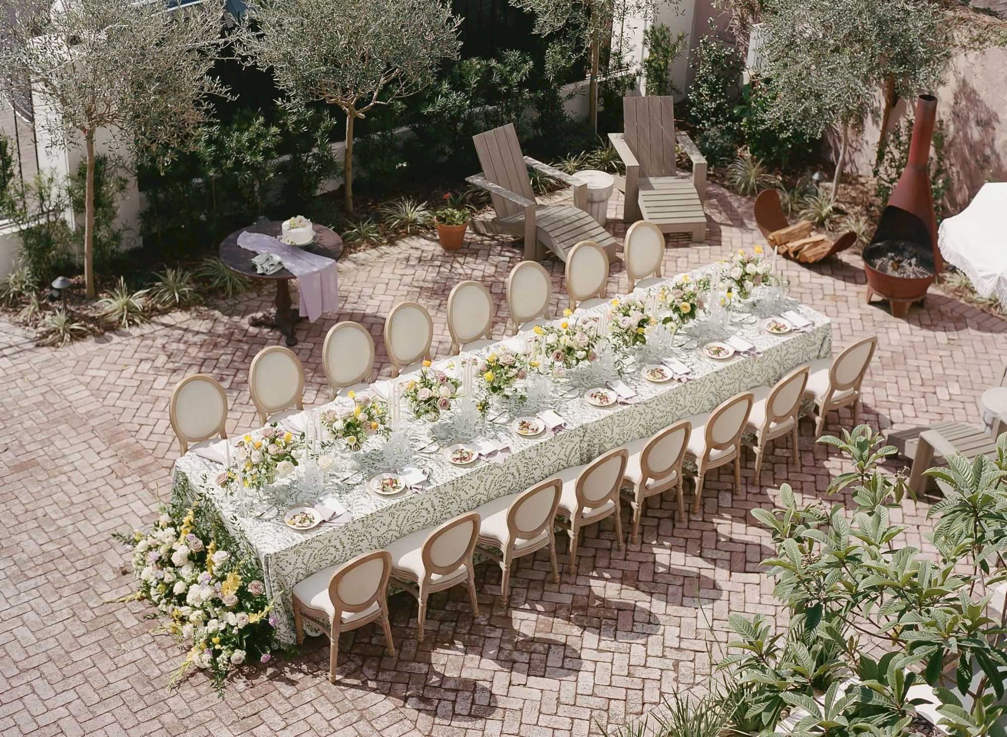 An outdoor dining setup with a long table, flower centerpieces, and 12 chairs, surrounded by greenery and garden furniture in a brick-paved area.