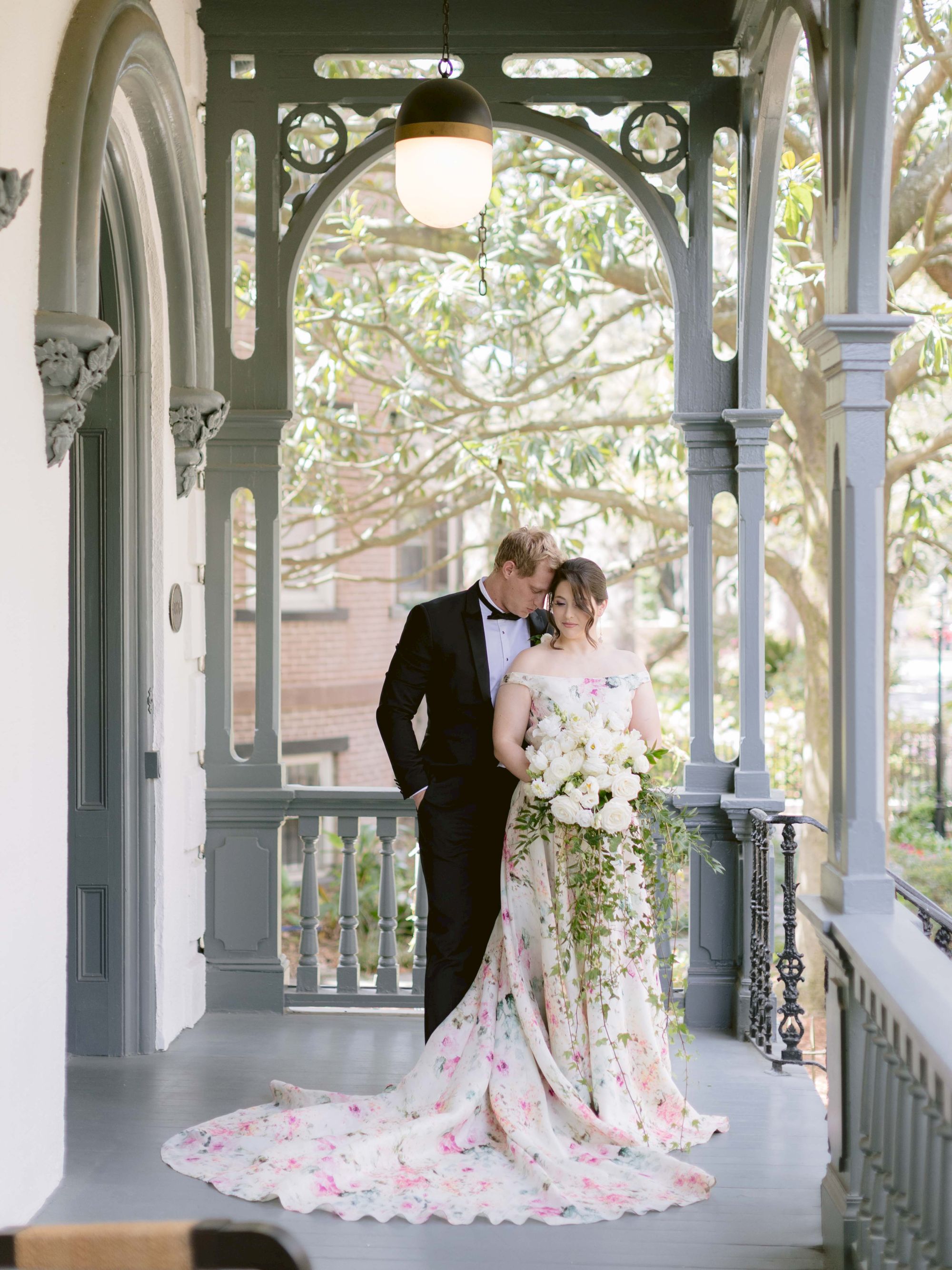 A couple dressed in wedding attire stands closely on a decorated porch with greenery in the background, sharing a tender moment.
