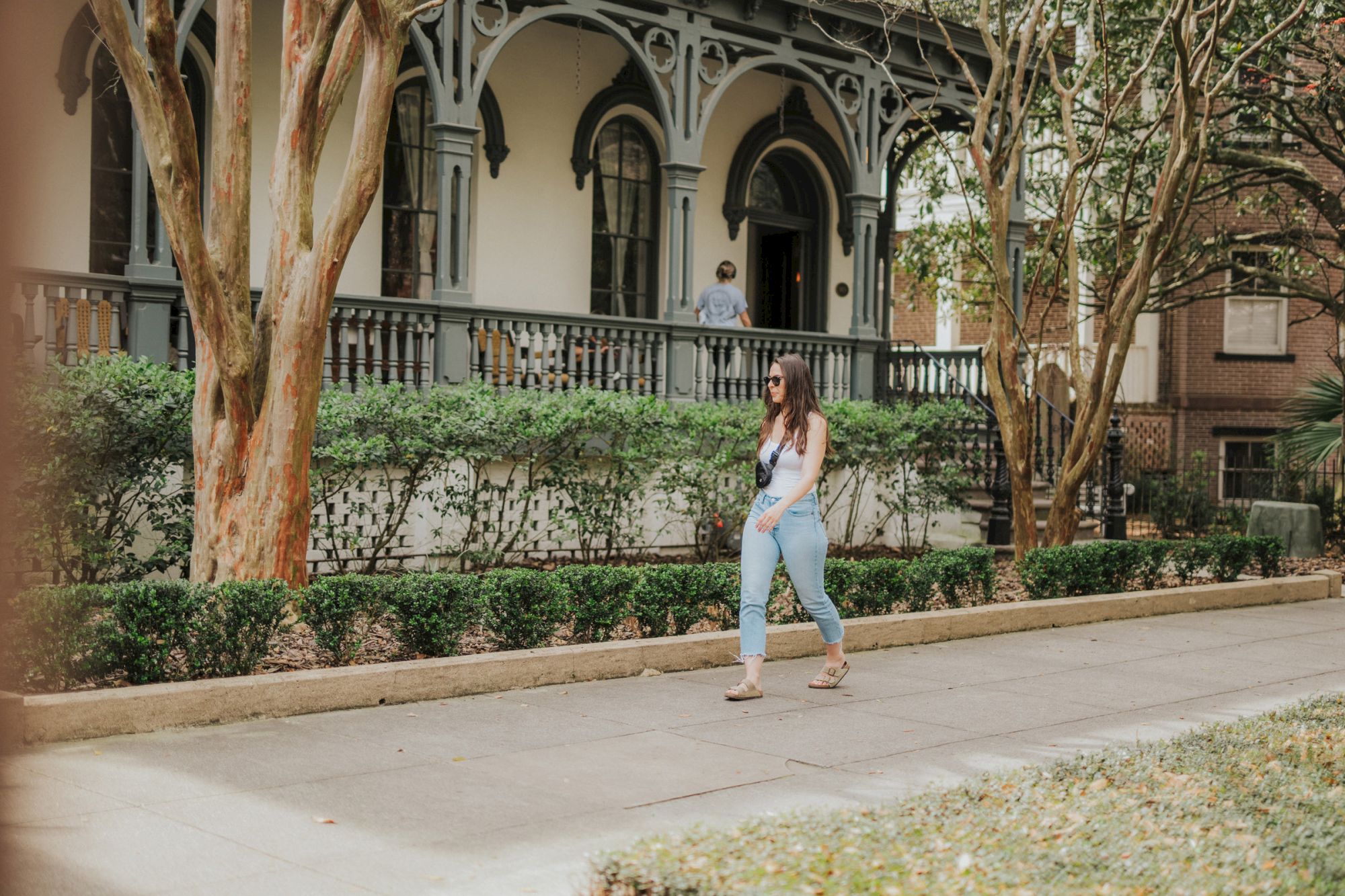 A woman is walking on a sidewalk in front of a house with ornate architectural features and surrounded by greenery.