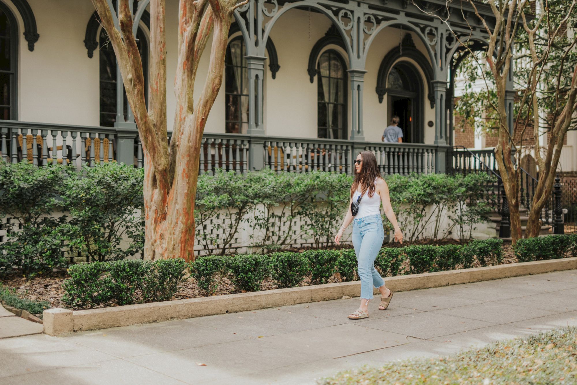 A woman is walking on a sidewalk in front of a building with a large porch and detailed architecture, surrounded by greenery, ending the sentence.