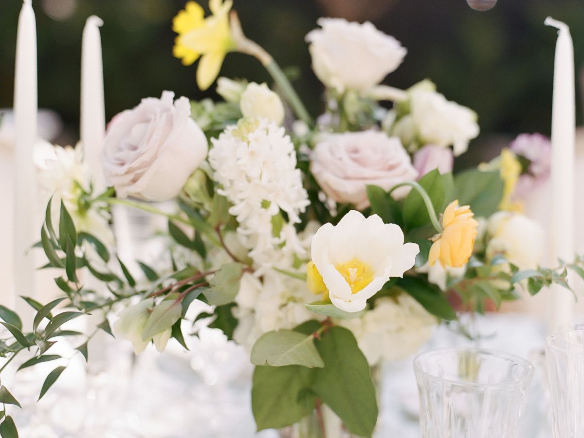 A floral centerpiece with yellow and white flowers on a table. The table setup includes candles, glassware, and white tablecloth.
