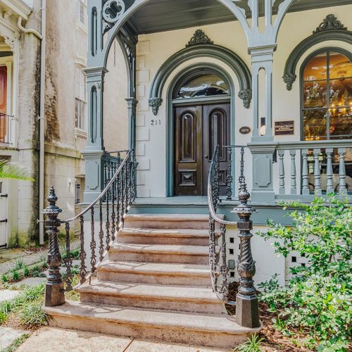 An ornate, vintage-style home with a wooden door, intricate iron railings, balcony, and arched windows surrounded by lush greenery.