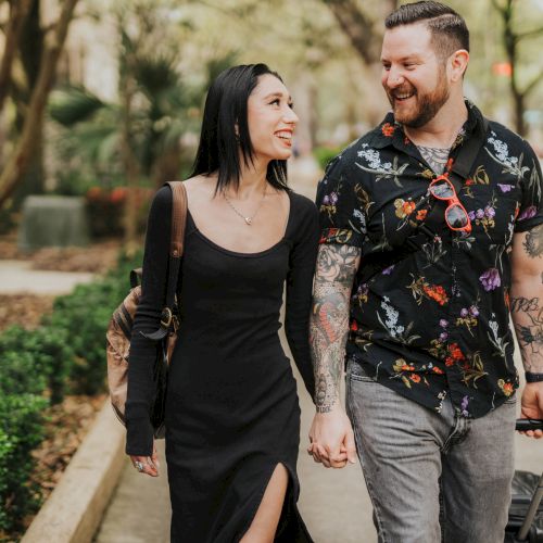 A couple is holding hands and walking along a path, smiling at each other. One is wearing a black dress, and the other has tattoos and a patterned shirt.