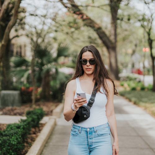 A woman is walking on a tree-lined sidewalk, looking at her phone while wearing sunglasses, a white tank top, and jeans.
