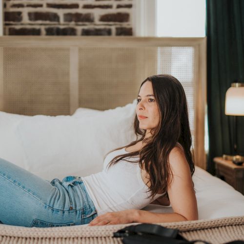 A woman in a white tank top and blue jeans is lying on a bed in a bedroom with a brick wall and lamp in the background, looking contemplative.