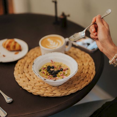A person is having a meal with a bowl of cereal topped with berries, a croissant, and a cup of coffee on a table with utensils.