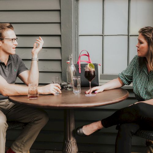 Two people sit at a round table outside, engaged in conversation, with drinks and a pink handbag on the table, ending the sentence.