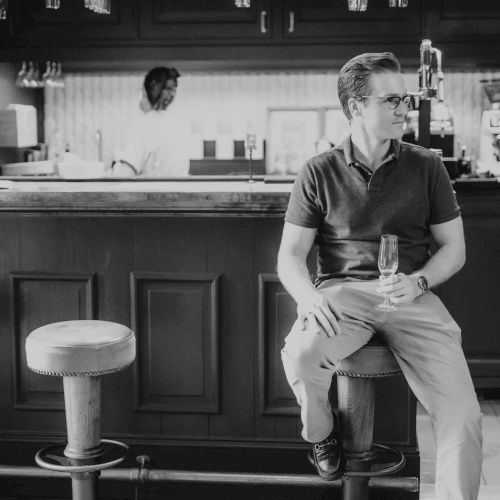 A person is sitting at a bar holding a drink, with a bartender in the background. The image is in black and white, giving it a classic feel.