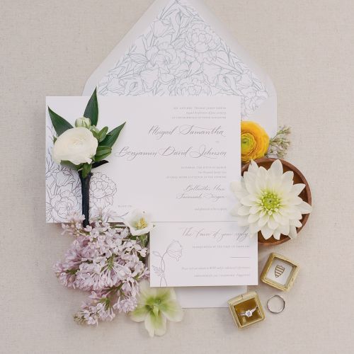 This image features wedding invitations, white flowers, a boutonnière, and two rings in boxes, all arranged on a beige background.