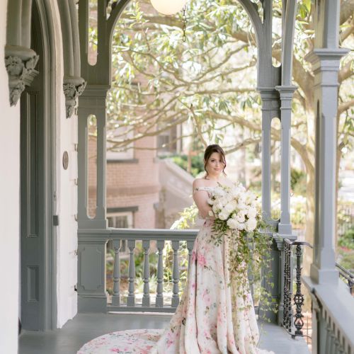 A woman in a floral gown stands on a beautifully decorated porch, holding a bouquet, with greenery and an elegant background.