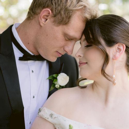 A couple dressed in wedding attire, with the groom in a black tuxedo and bow tie, and the bride in an off-shoulder dress, sharing an intimate moment.