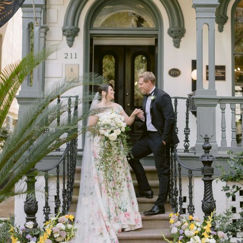 A bride and groom stand on the steps of a decorated building, surrounded by flowers, holding hands and gazing at each other lovingly.