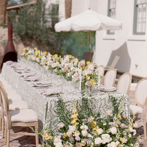 An elegant outdoor dining setup features a long table with floral arrangements, fine tableware, and an umbrella, surrounded by stylish chairs.