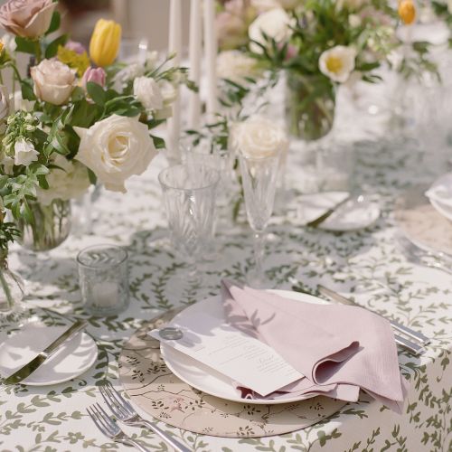 A table setup with floral decorations, candles, cutlery, glassware, and a plate with a folded pink napkin on a white tablecloth with green patterns.