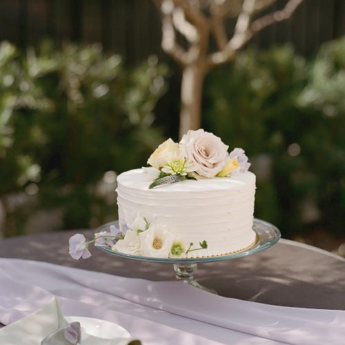 A white frosted cake with floral decorations is on a glass cake stand, placed on a table with a folded napkin and cutlery, set outdoors.