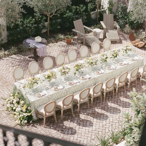 An elegant outdoor dining setup with a long, decorated table featuring floral arrangements and cushioned chairs in a courtyard with greenery.