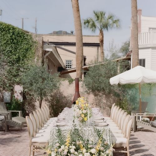 An outdoor dining setup with a long table, floral centerpiece, and chairs around it. The setting has palm trees and a surrounding patio area, ending the sentence.