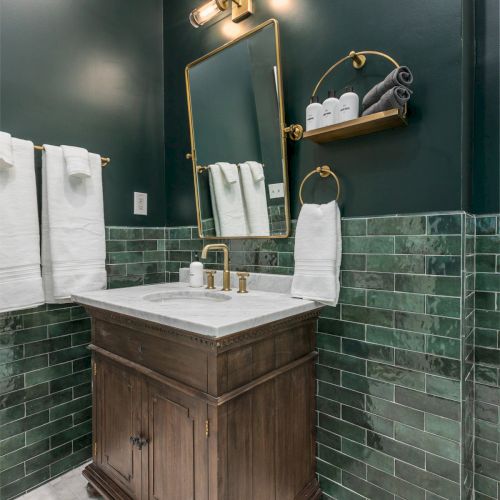 A small bathroom with dark green tiles, a wooden vanity with a white countertop, a mirror, gold fixtures, and neatly folded towels on a shelf.