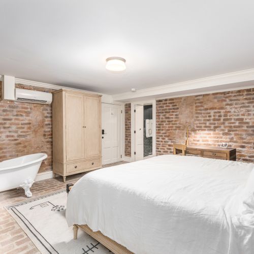 A bedroom with a brick wall, white bedding, a freestanding bathtub, wooden wardrobe, desk, and plants, under ceiling lighting ends the sentence.