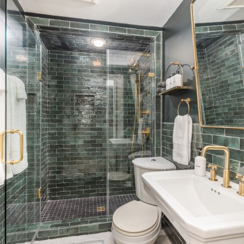 A modern bathroom features a glass-enclosed shower with dark green tiles, a white toilet, a pedestal sink, a large mirror, and gold fixtures.