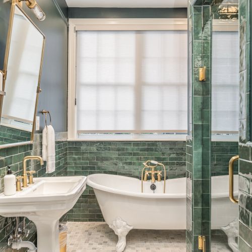 A bathroom with green tiles, a white clawfoot bathtub, a pedestal sink, a large mirror, and glass shower, illuminated by natural light.