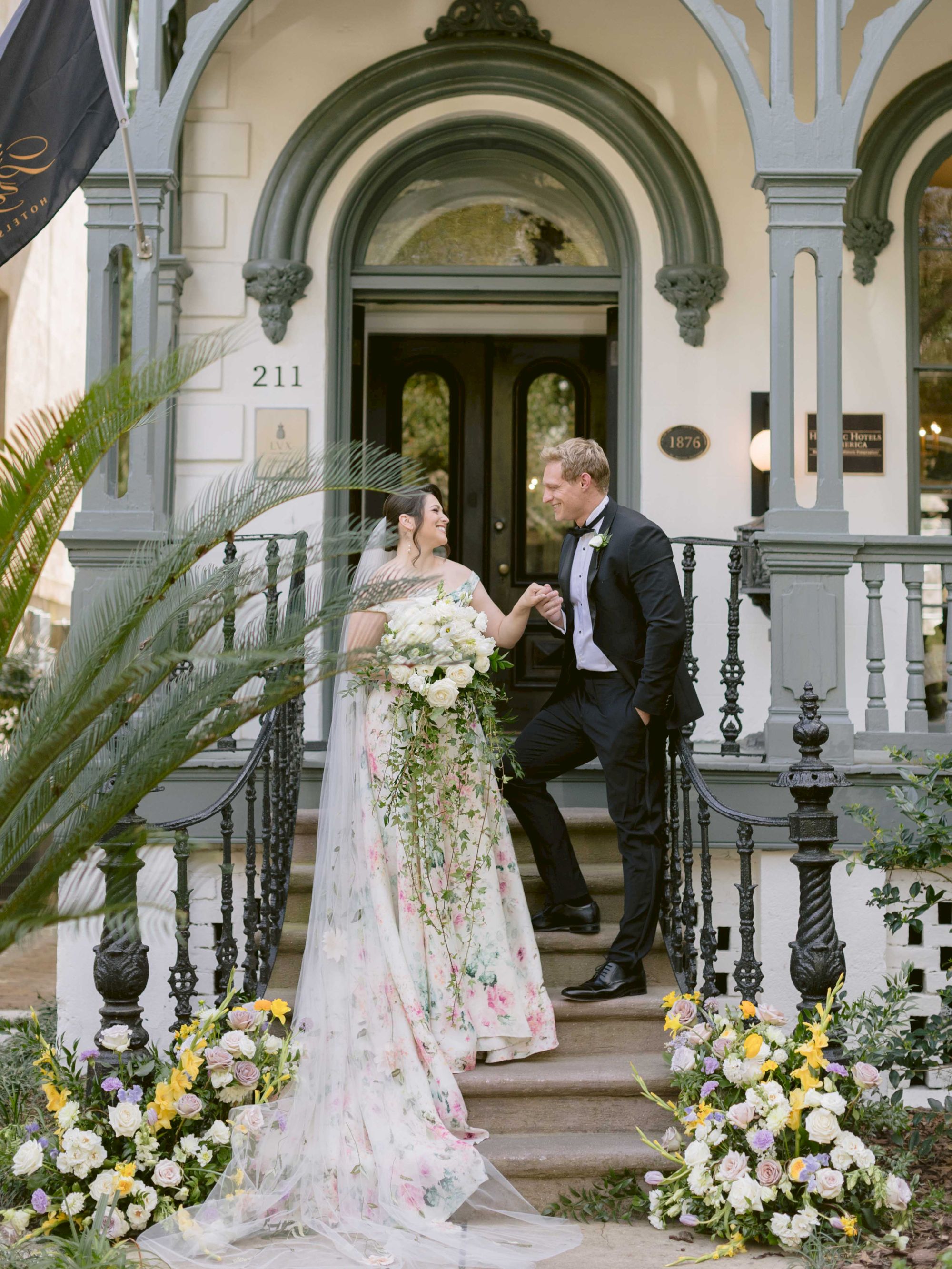 A bride and groom pose on the steps of a decorative building, surrounded by greenery and flowers, creating a picturesque wedding scene.