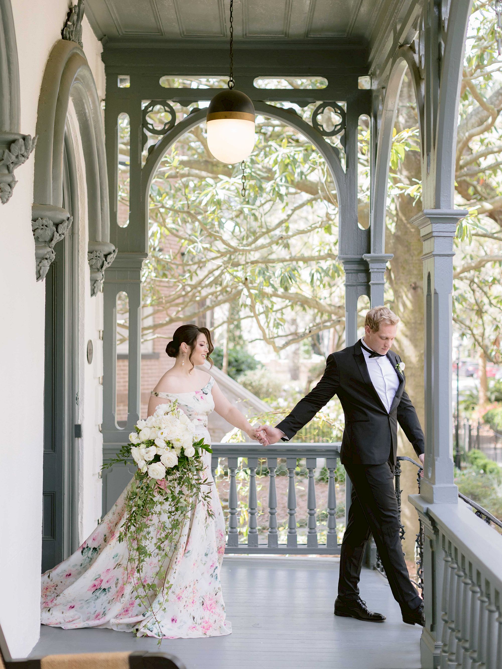 A couple in wedding attire is holding hands on a decorated porch, surrounded by greenery. The bride is in a floral gown, and the groom is in a suit.
