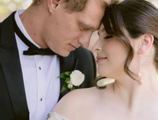 A couple dressed in wedding attire share an intimate moment, their foreheads touching, with a lush, floral background.