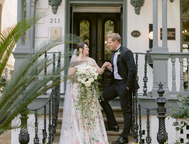 A couple in wedding attire is posed on the steps of a decorated building, surrounded by flowers and greenery, creating a romantic atmosphere.