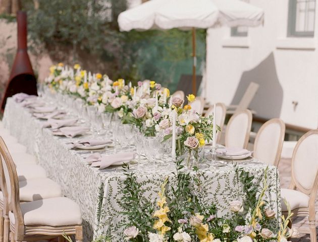 An outdoor dining table is elegantly set with floral arrangements and white tableware. The setting includes chairs and a white umbrella for shade.