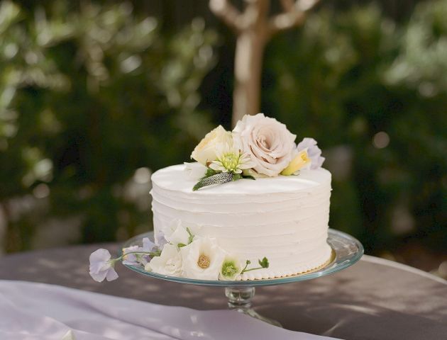 The image shows a white cake decorated with flowers on a glass stand, set on an outdoor table with greenery in the background.