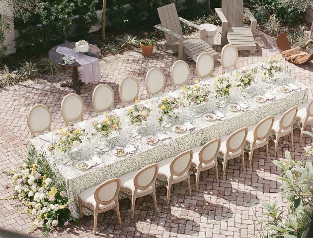 The image shows an elegantly set outdoor dining table with floral arrangements, surrounded by chairs, situated on a brick patio.