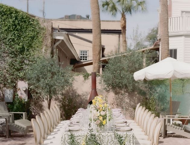 An elegant outdoor dining setup with a long table, chairs, and floral arrangements, situated in a courtyard with palm trees and surrounding greenery.